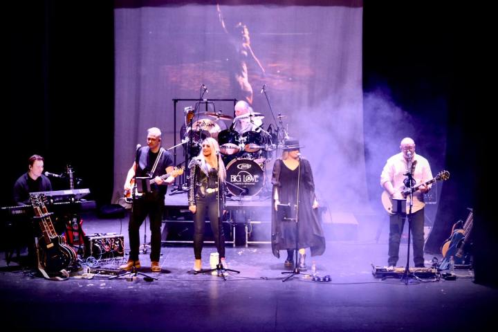 Five-piece band performing on stage with guitars, keyboard, and drums. Purple lighting and fog add ambiance.