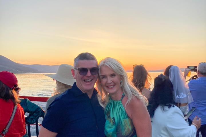 Couple smiling on a boat deck at sunset with people and mountains in background.